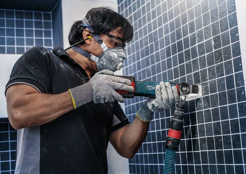 Une personne porte des vêtements de protection et utilise un outil électrique pour travailler des carreaux sur un mur.