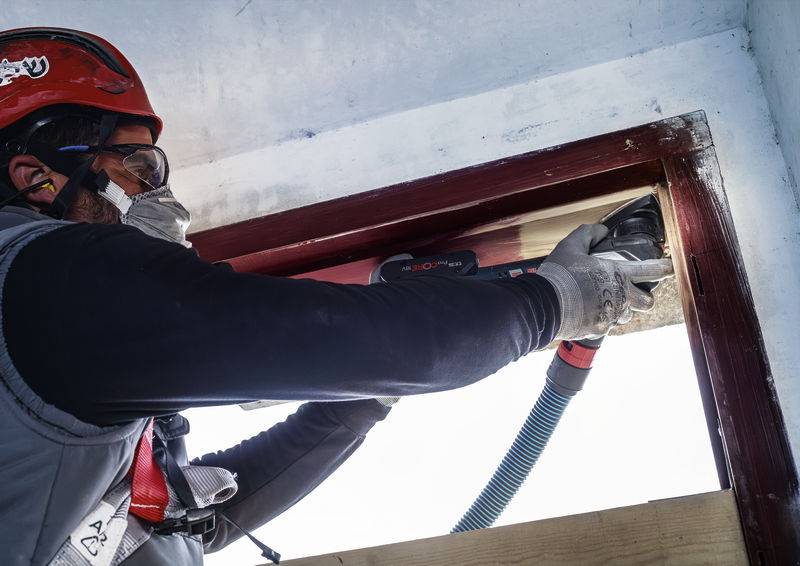 Une personne portant un équipement de protection utilise un outil électrique pour travailler sur un encadrement de porte, probablement dans le cadre d'une rénovation ou d'une réparation.