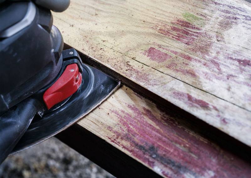 Une ponceuse lisse la surface rugueuse d'une table en bois. Des résidus rouges sont visibles, ce qui indique que de la peinture a été poncée.