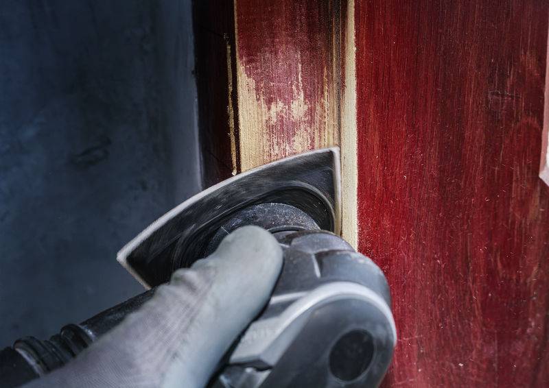 Gros plan sur une personne qui retire de la peinture rouge d'une surface en bois à l'aide d'un outil de ponçage. Un gant de protection est visible.