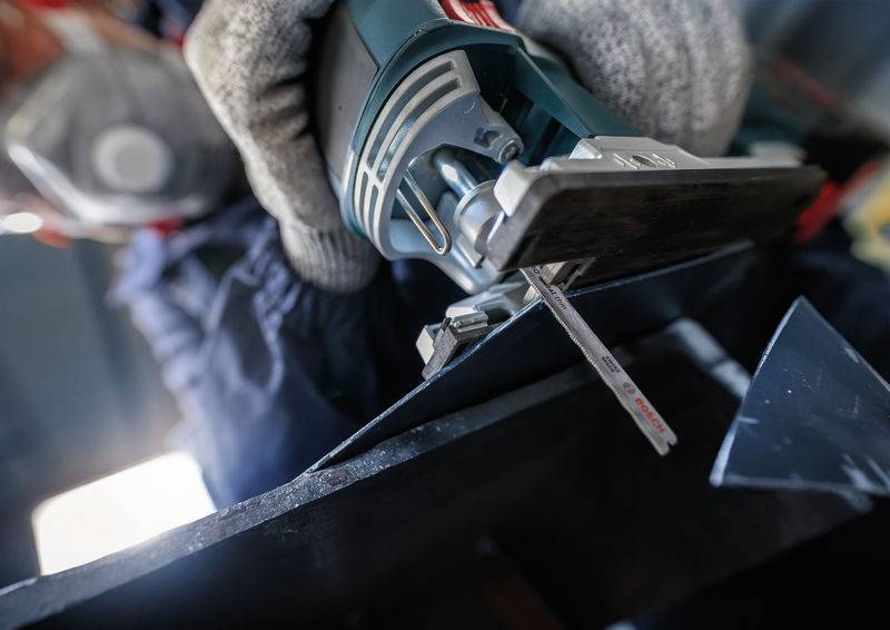 Une personne coupe un morceau de métal à l'aide d'une scie sauteuse. Elle porte des gants de travail et des protections auditives, concentrée sur sa tâche.
