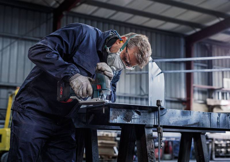 Un homme portant des lunettes de protection et un masque coupe du métal à l'aide d'une scie électrique dans un environnement industriel.