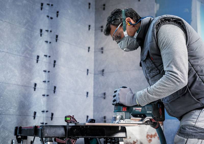 Un artisan portant des lunettes de protection et un masque utilise un outil électrique pour travailler un matériau sur un établi.