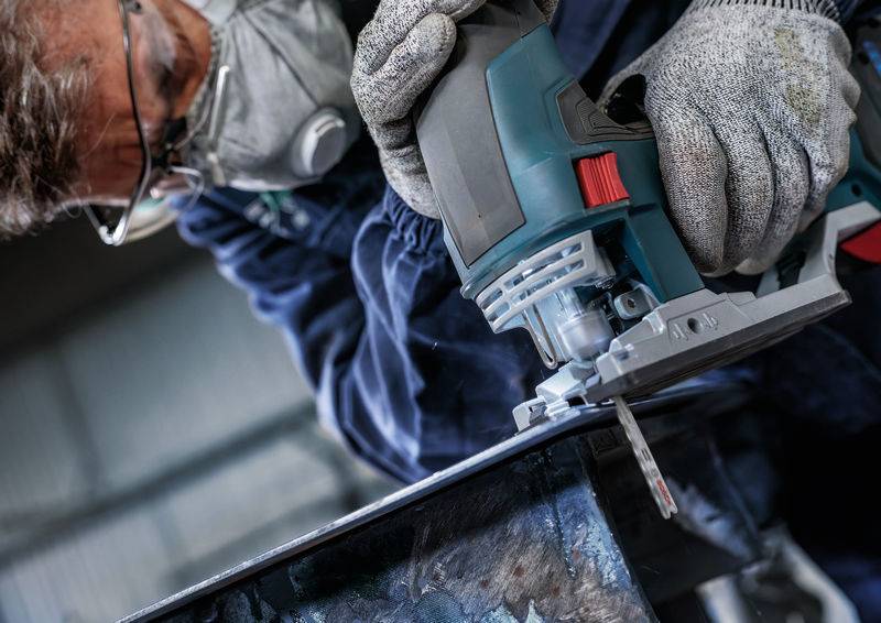 Une personne vêtue d'un équipement de protection et portant un masque utilise une scie sauteuse électrique pour couper un morceau de métal.