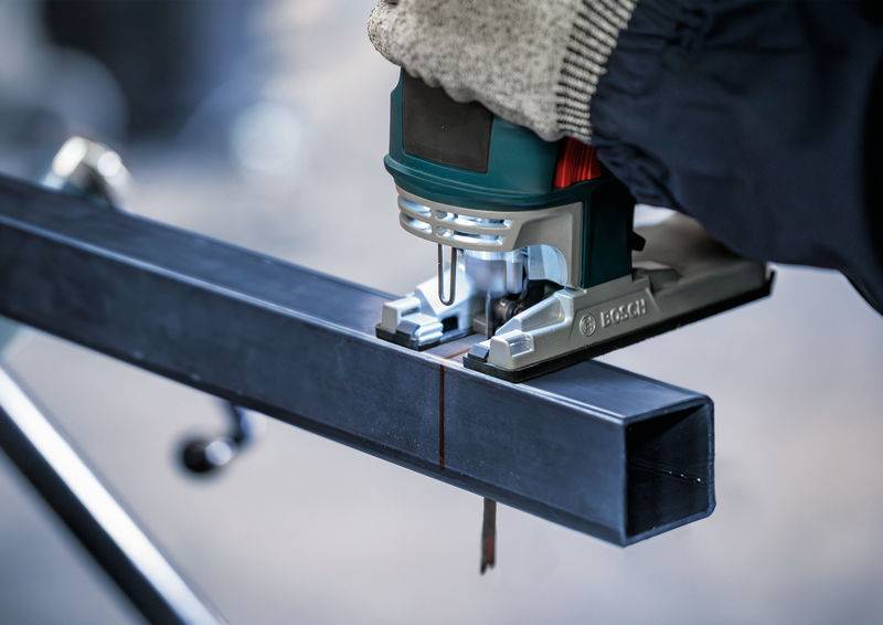 Une personne utilise une scie sauteuse pour couper un profilé de rail métallique le long d'une ligne marquée. Elle porte des gants.