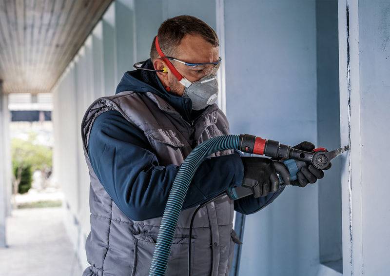 Une personne en tenue de protection utilise un outil électrique sur un mur. Elle porte des lunettes de protection, des protections auditives et un masque.