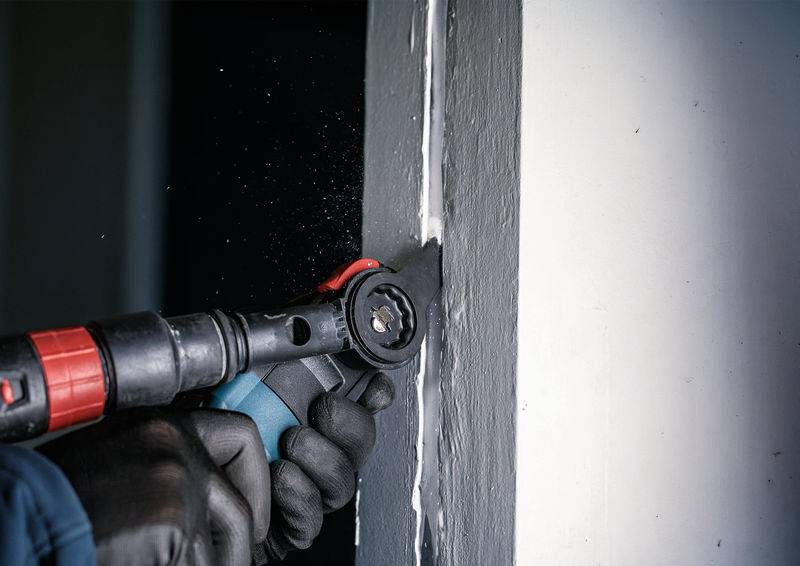 Une personne découpe du matériau d'étanchéité dans un joint de mur blanc à l'aide d'un outil oscillant. Elle porte des gants noirs.