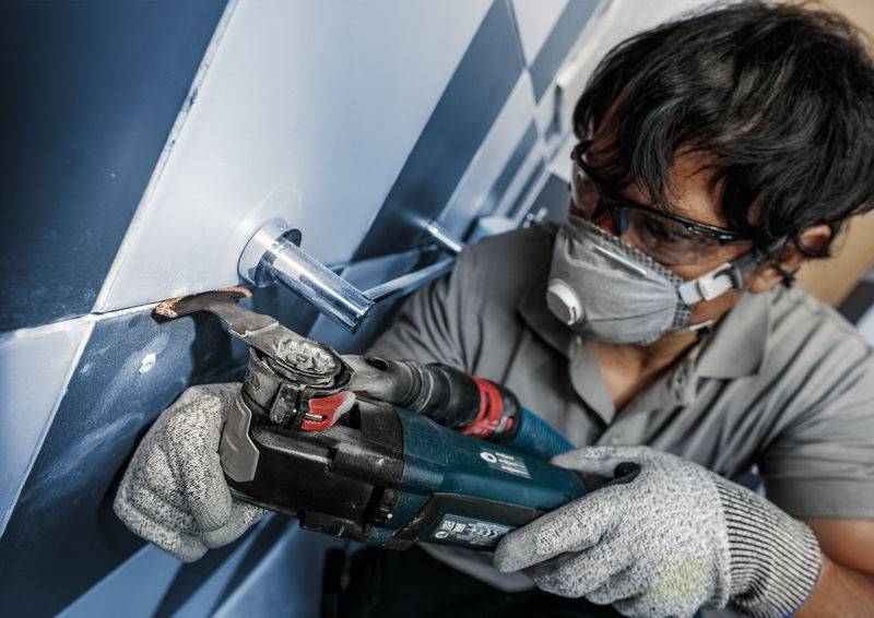 Une personne porte des lunettes de protection et un masque respiratoire, utilisant un outil électrique pour travailler des carreaux sur un mur.