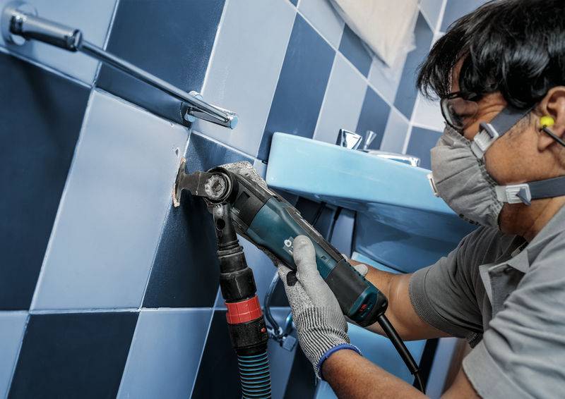 Une personne porte un masque de protection et des protections auditives, et utilise une meuleuse électrique pour travailler des carrelages dans une salle de bain.