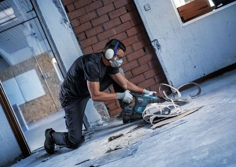 Un ouvrier portant des protections auditives et des lunettes de sécurité travaille avec un outil électrique sur un sol dans un bâtiment en construction.