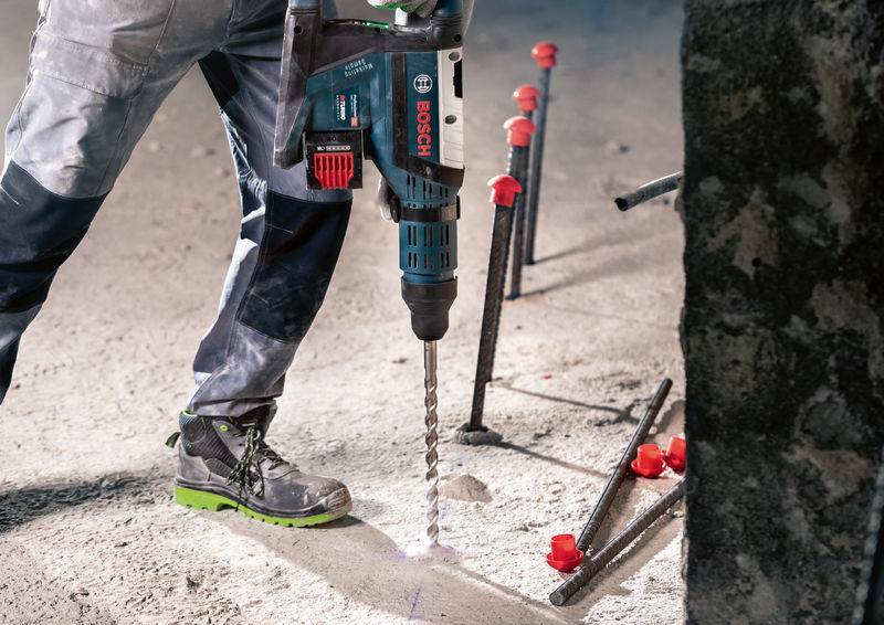 Une personne fore un sol en béton avec un marteau-piqueur Bosch, entourée de barres métalliques apparentes et de couvertures en plastique rouge.