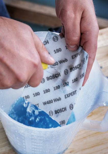 Une personne trempe un papier de verre Bosch portant l'inscription 'EXPERT W480' dans un bécher contenant un liquide bleu.