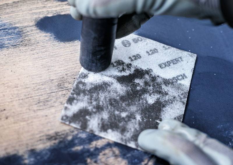 Une personne ponce une surface en bois avec du papier de verre. De la poussière se répand sur la surface.