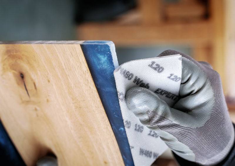 Une personne ponce le bord d'un morceau de bois avec un bloc de papier de verre. Elle porte des gants.