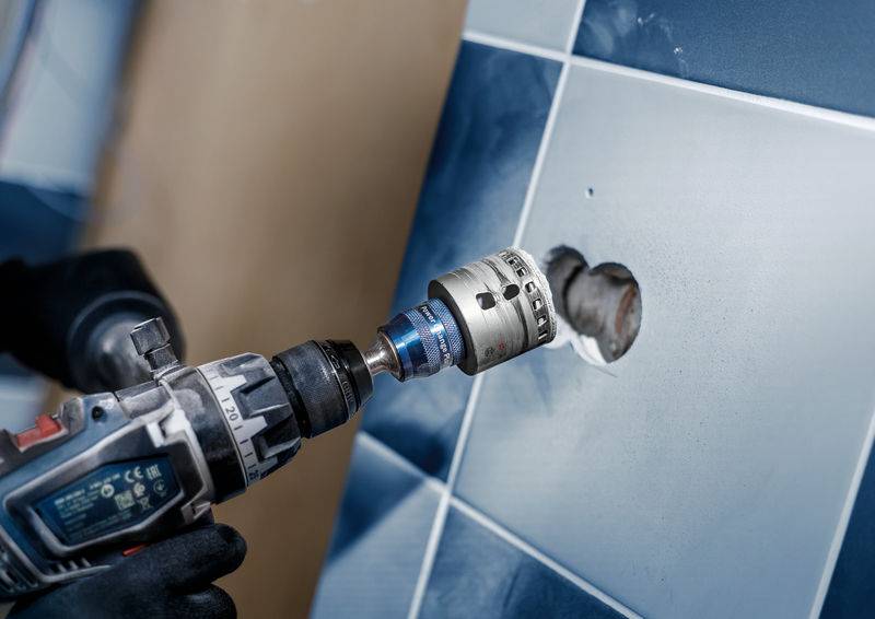 Une personne fore un trou rond dans des carreaux bleus sur un mur à l'aide d'une perceuse.