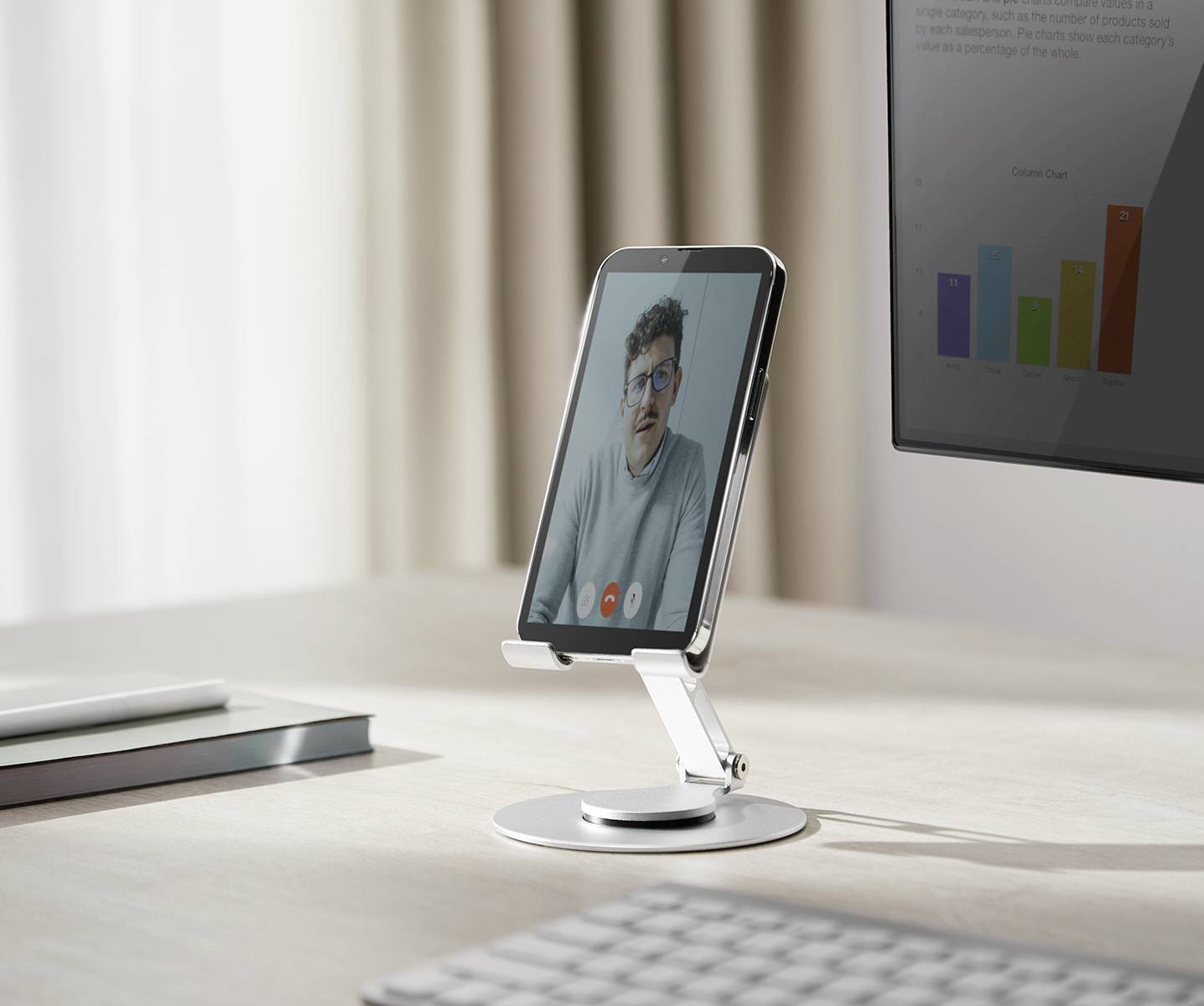 Un smartphone sur un support affiche une visioconférence avec une personne. À côté se trouve un écran qui montre un graphique à barres.