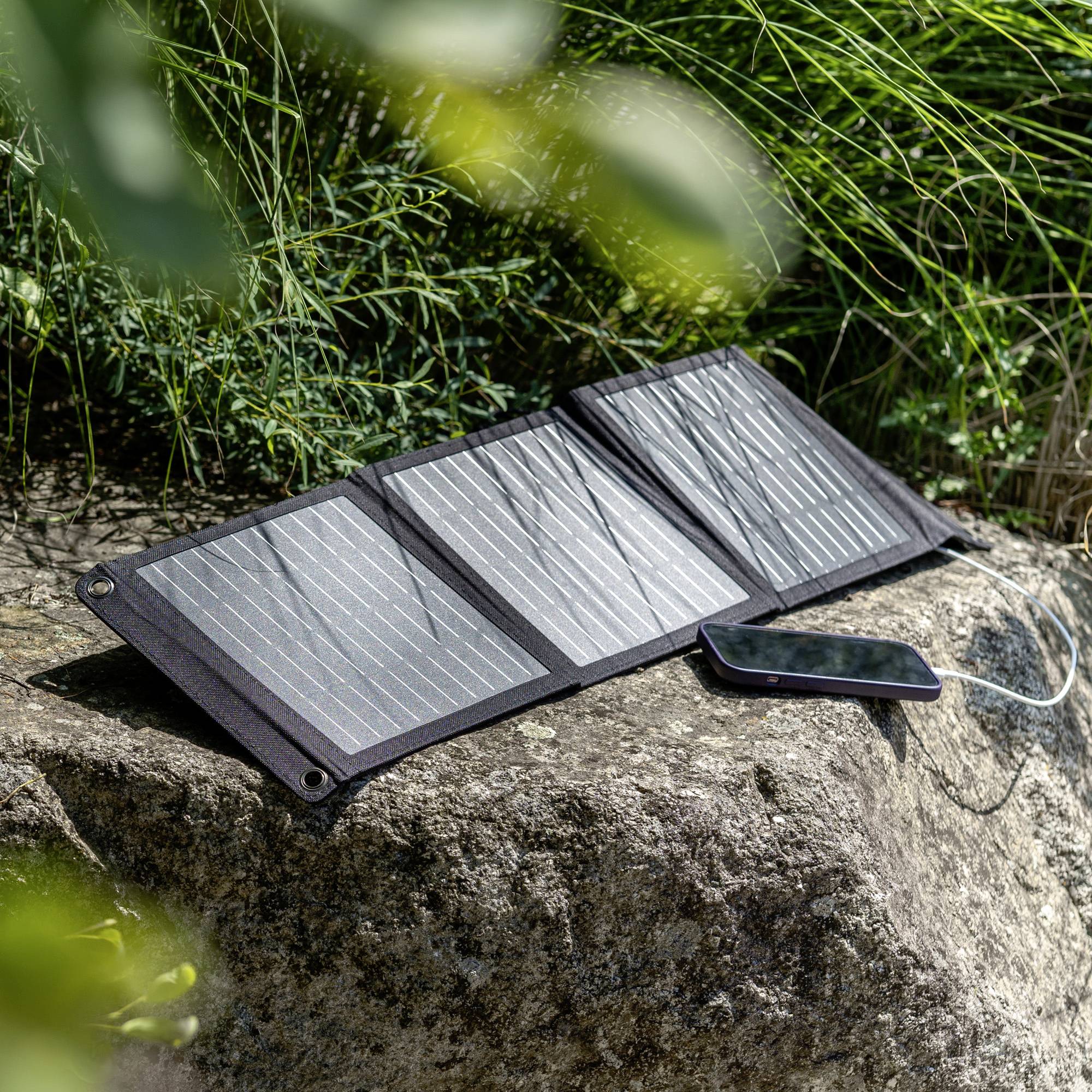Un panneau solaire pliable est posé sur un rocher au soleil. Un smartphone est connecté et en train de se charger via un câble.