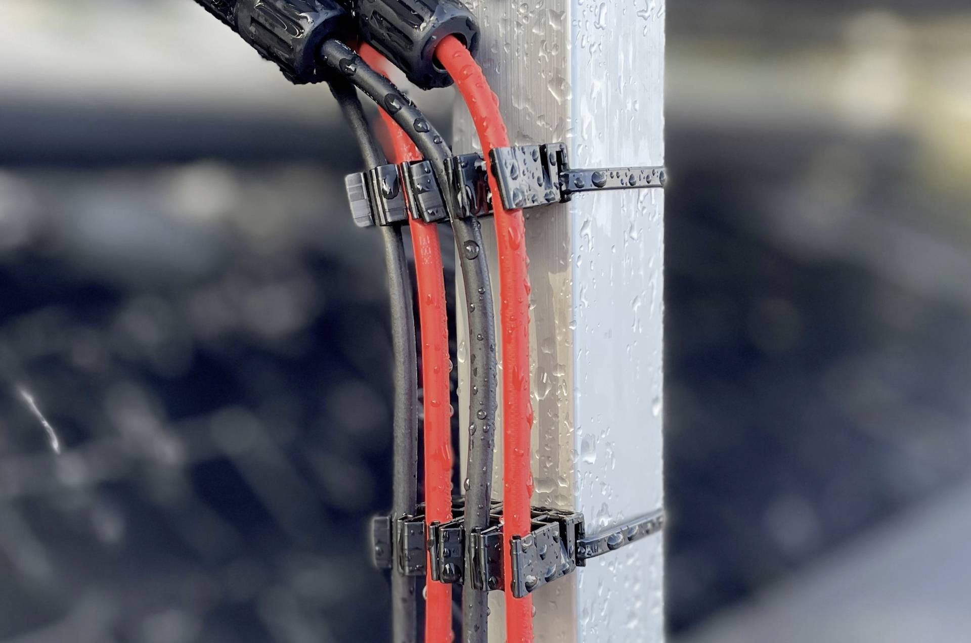 Des câbles solaires rouges sont fixés avec des clips sur un profilé métallique, avec en arrière-plan une vue floue de panneaux solaires.
