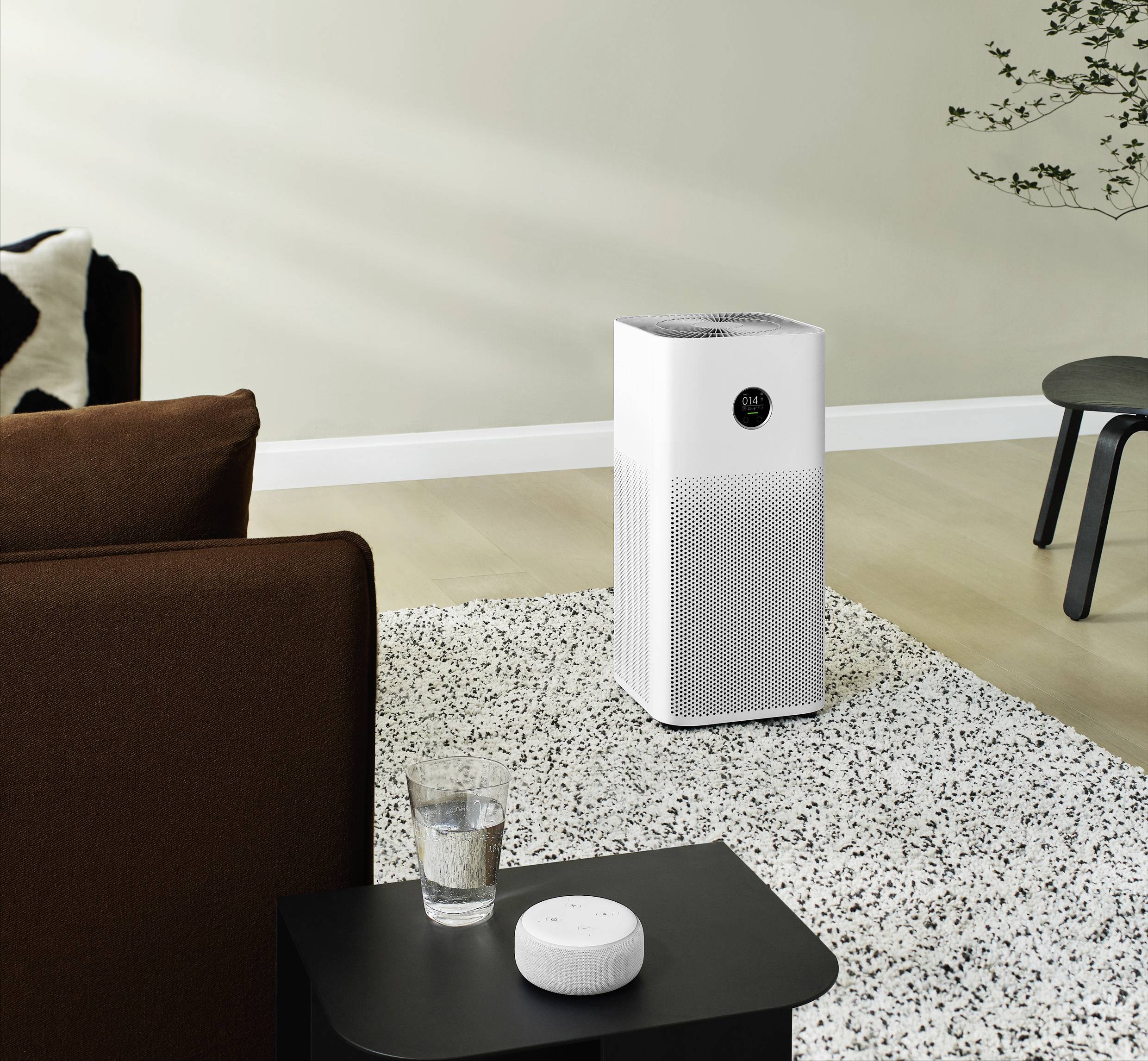 Un purificateur d'air est posé sur un tapis clair dans un salon moderne. Une petite table avec un verre d'eau se trouve à côté.