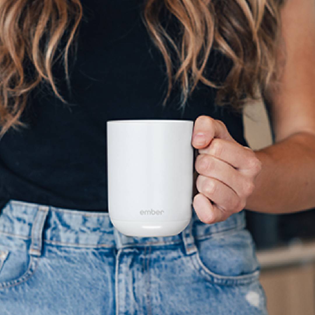 Une personne tient une tasse blanche dans sa main droite, porte un haut noir et un jean bleu.