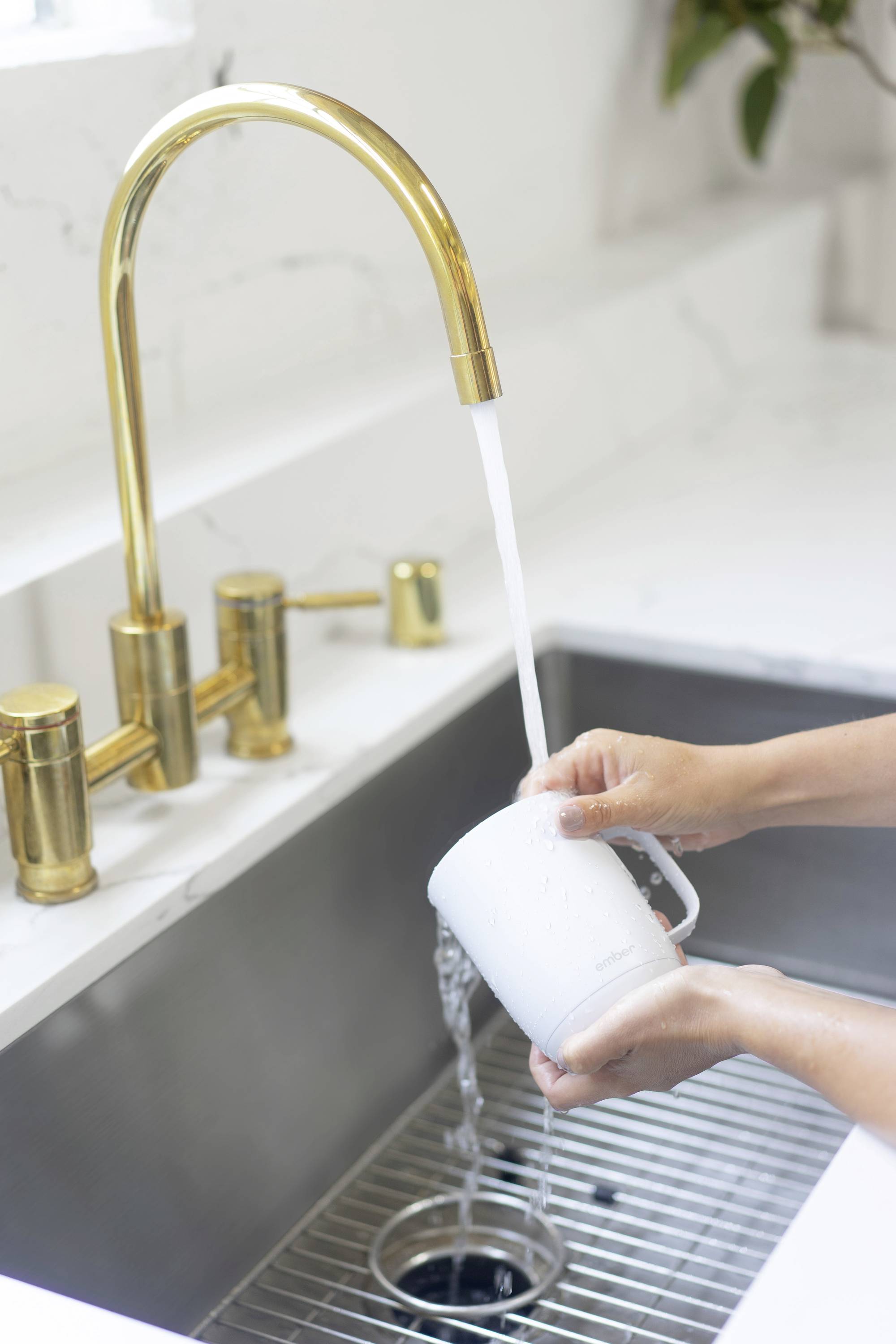 Des mains rincent une tasse blanche sous l'eau courante dans un lavabo moderne avec un robinet doré.