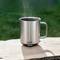 Tasse en métal avec une boisson fumante sur une table en bois à l'extérieur. La tasse a une anse et la marque 'ember' est imprimée dessus.