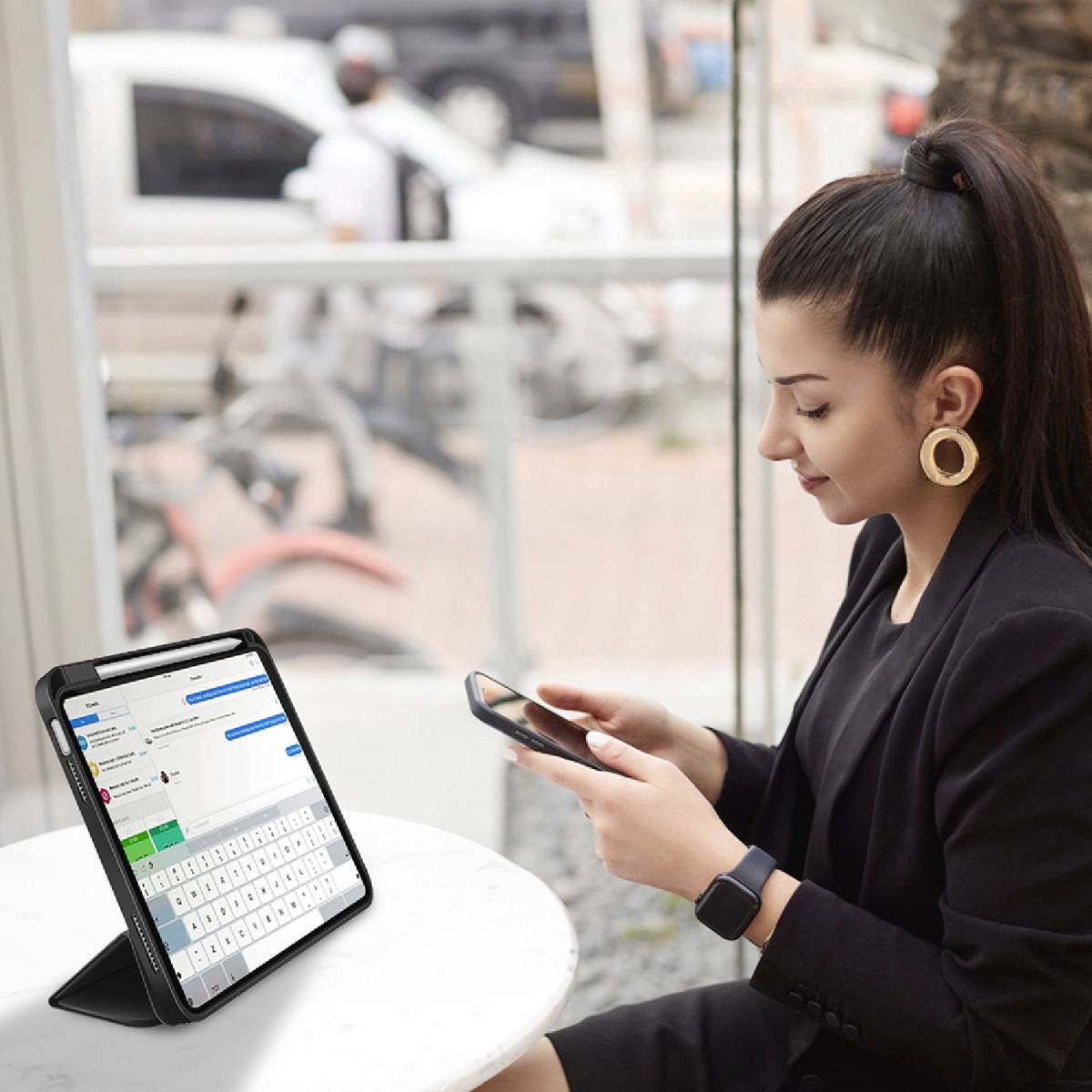 Une femme est assise dans un café et regarde son smartphone. À côté d'elle, une tablette est posée sur une table et affiche une application de chat.