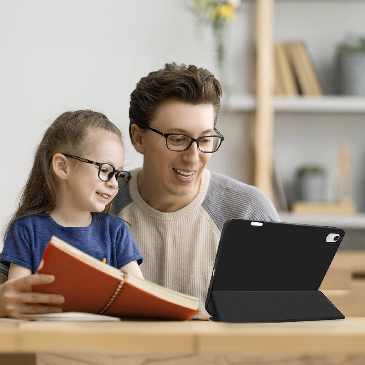 Un adulte souriant est assis à une table avec un enfant, tous deux portant des lunettes et regardant une tablette posée devant eux. Des livres et une plante sont visibles en arrière-plan.