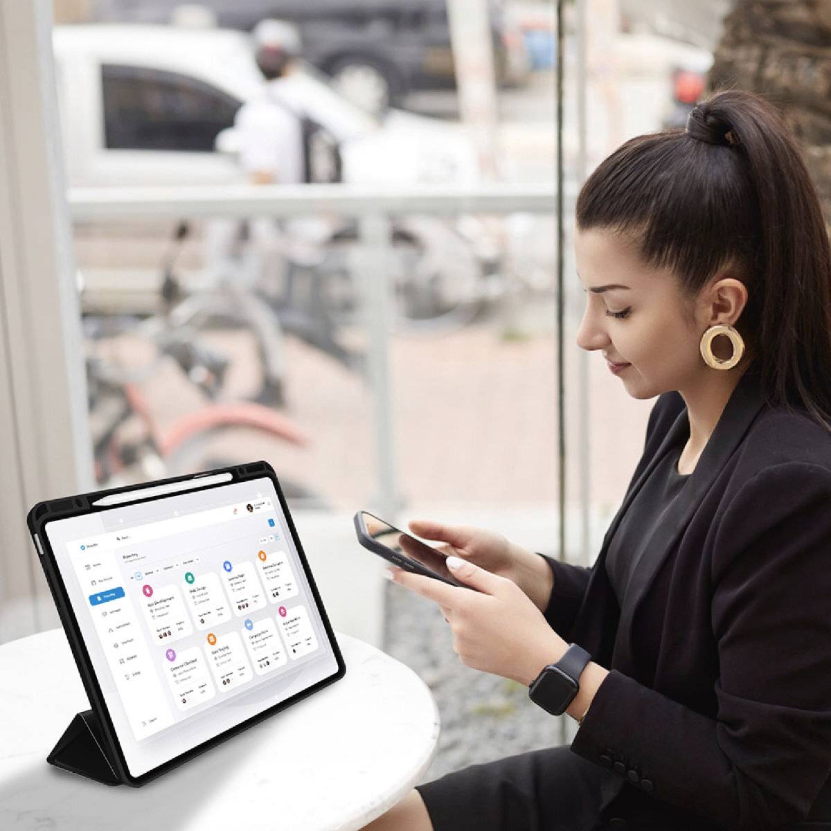 Femme assise à une table, tenant un smartphone, un tablet ouvert sur un calendrier à côté d'elle. Elle a l'air concentrée.