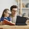 Un adulte et un enfant sont assis à table, rient et regardent une tablette, tandis que l'enfant tient un livre.