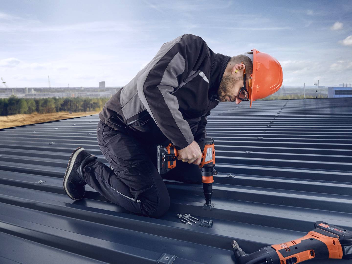 Un ouvrier portant un casque de protection orange et des lunettes est agenouillé sur un toit métallique et utilise une perceuse. Un paysage est visible en arrière-plan.