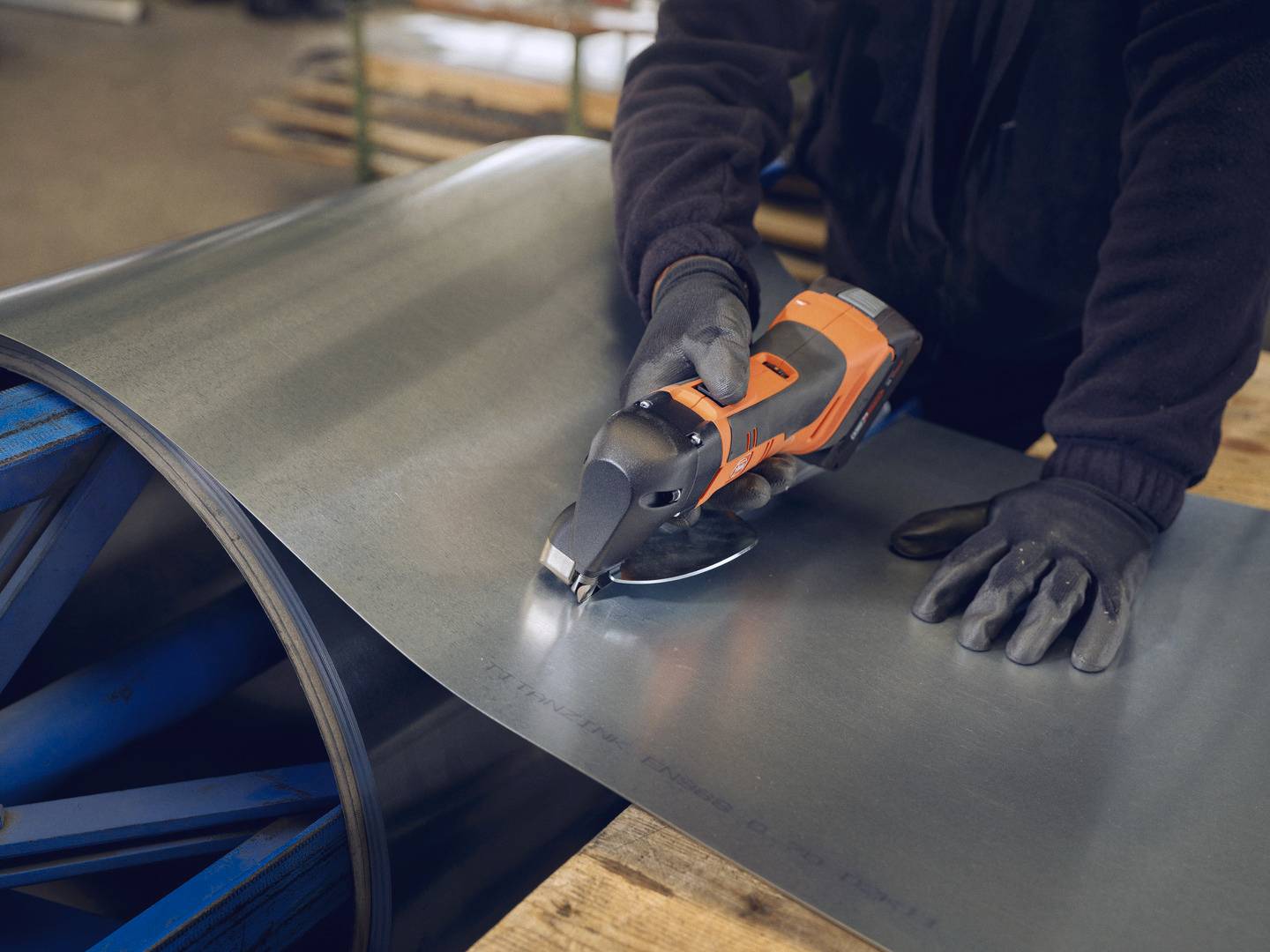 Une personne coupe une tôle métallique avec un outil électrique sur un établi. Elle porte des gants de protection.