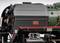 Gros plan d'une partie d'une locomotive à vapeur, révélant des détails techniques et une bande rouge sur du métal noir. Un symbole électrique est visible à gauche.