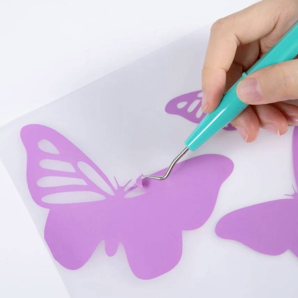 Une main tient un outil et retire une partie d'un design de papillon lilas à partir d'un vinyle sur un tapis transparent.