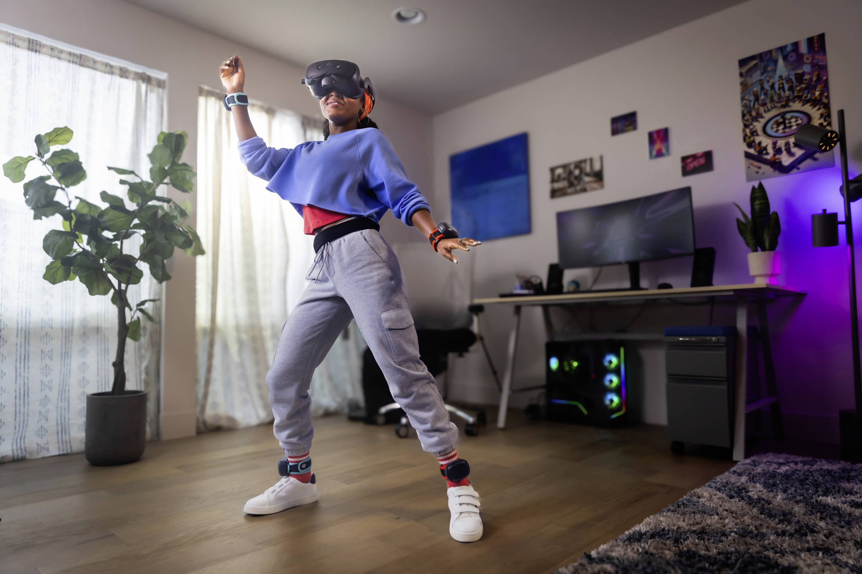 Une personne danse dans un espace moderne équipée d'un casque de réalité virtuelle, entourée d'un ordinateur et de plantes. Un éclairage vif et des vêtements décontractés complètent la scène.