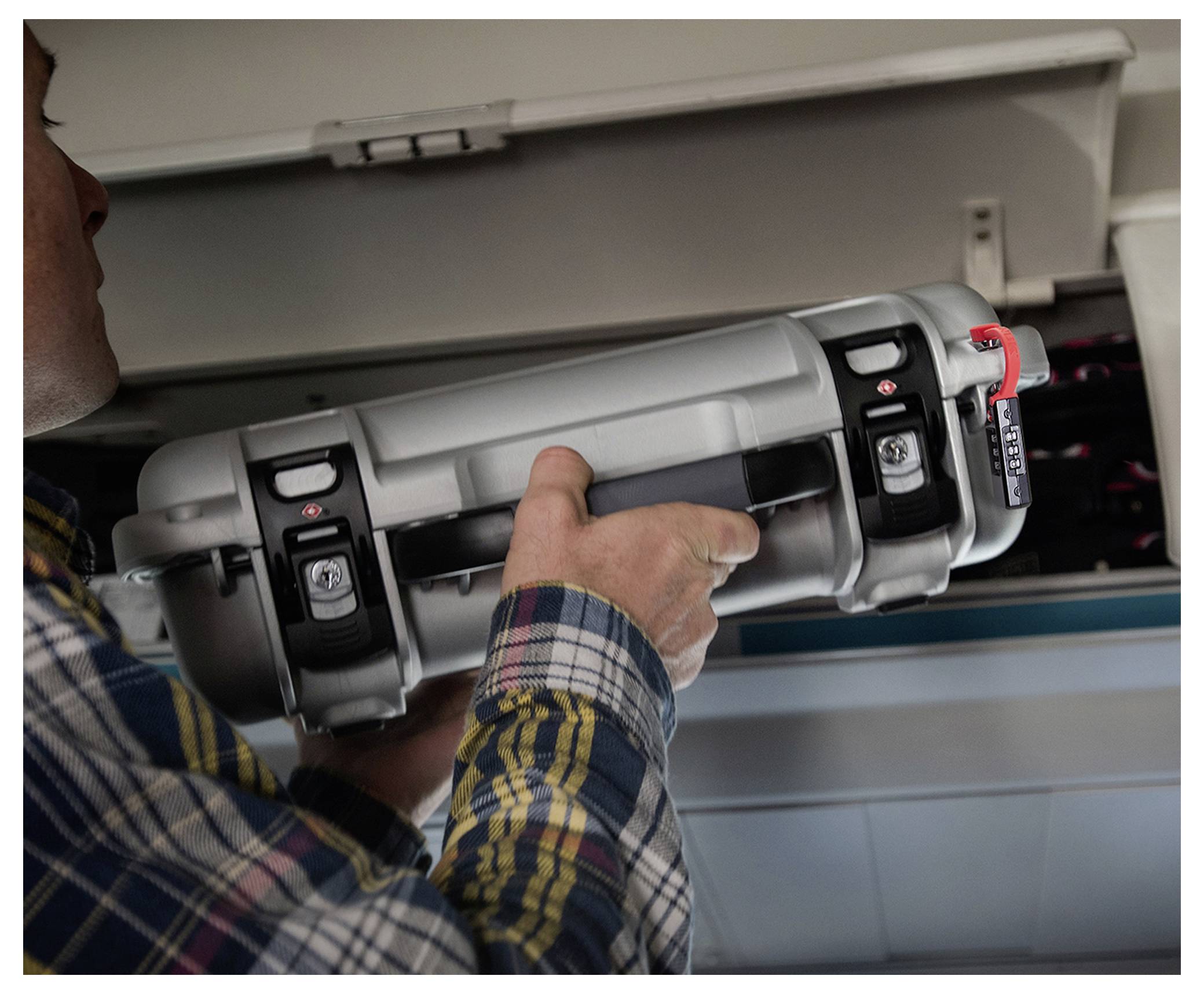 Une personne range une valise rigide grise dans un compartiment à bagages d'avion. La valise est équipée d'une fermeture métallique et d'un cadenas à code.