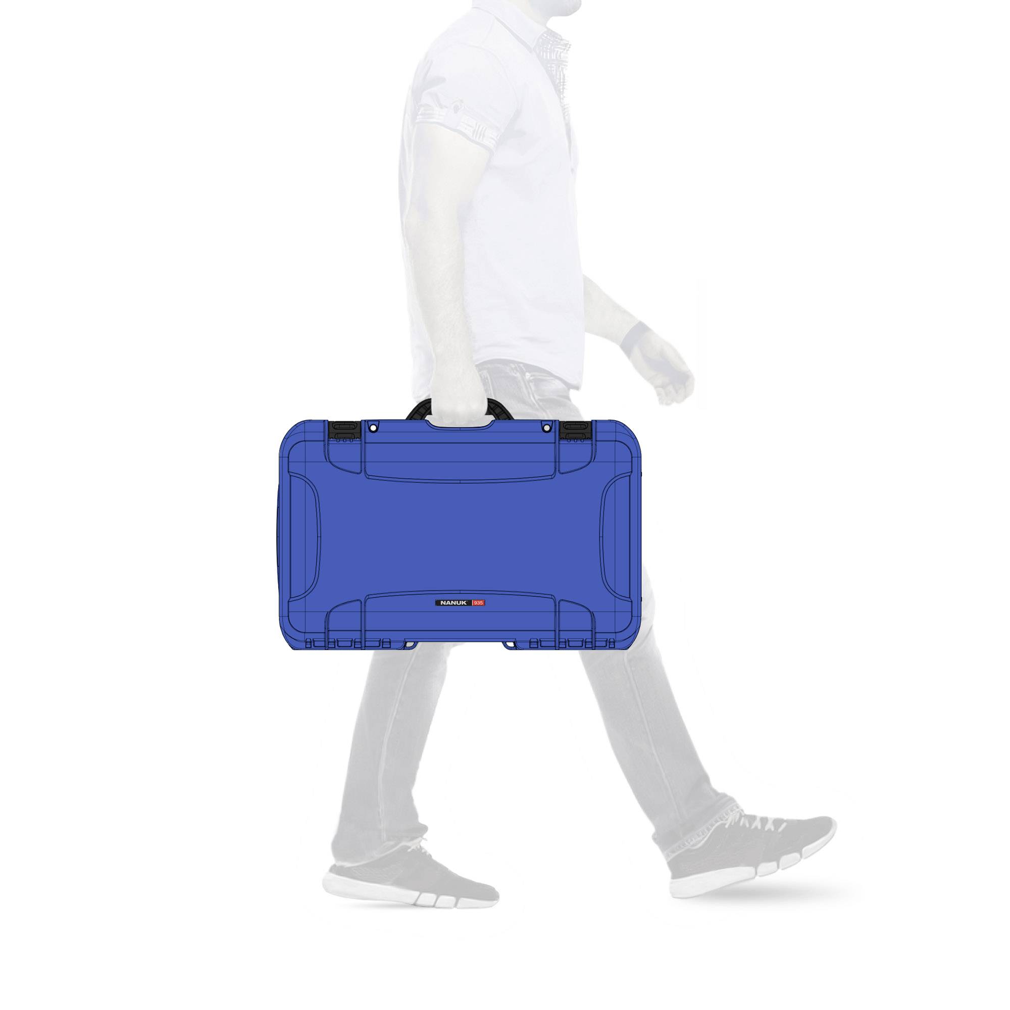 Un homme portant une chemise blanche et un jean transporte une valise bleue et marche devant un fond neutre.