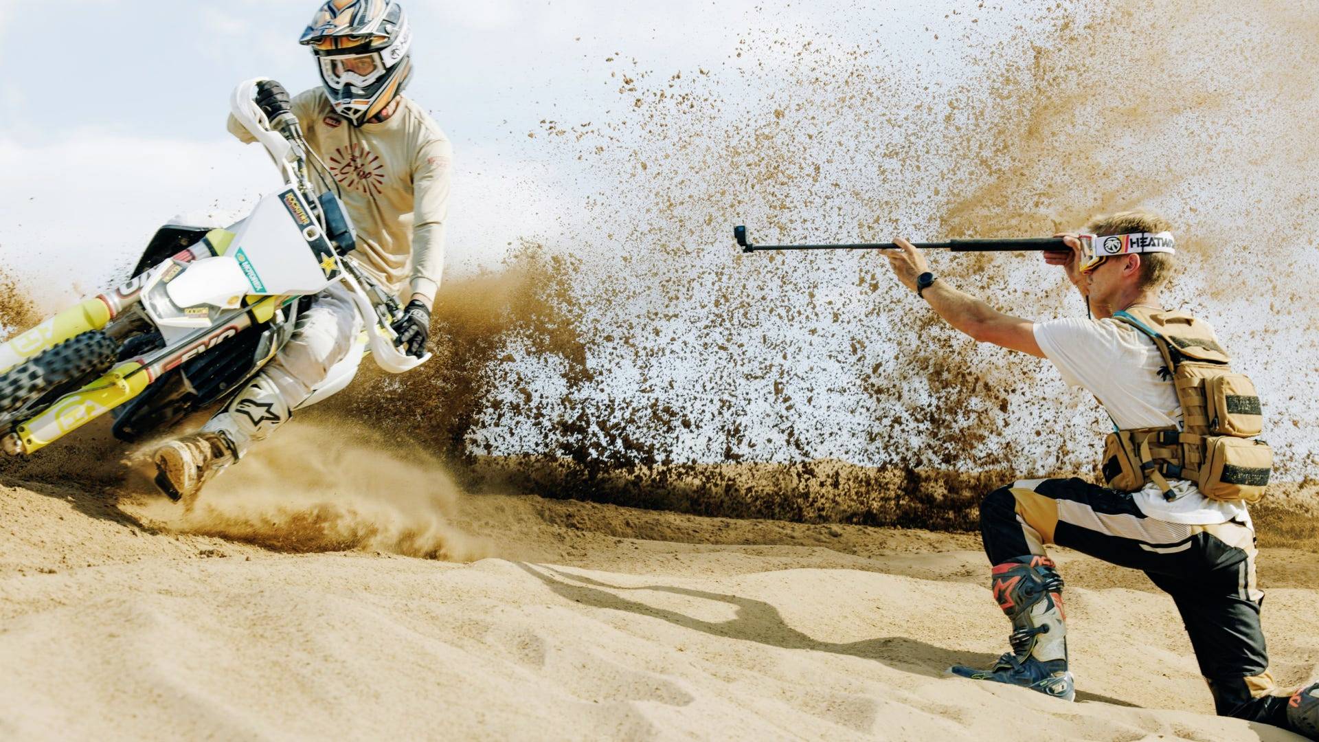 Un motard traverse un terrain sablonneux, soulevant un nuage de poussière, tandis qu'un photographe le capture avec une caméra d'action pour obtenir un cliché sportif dynamique.
