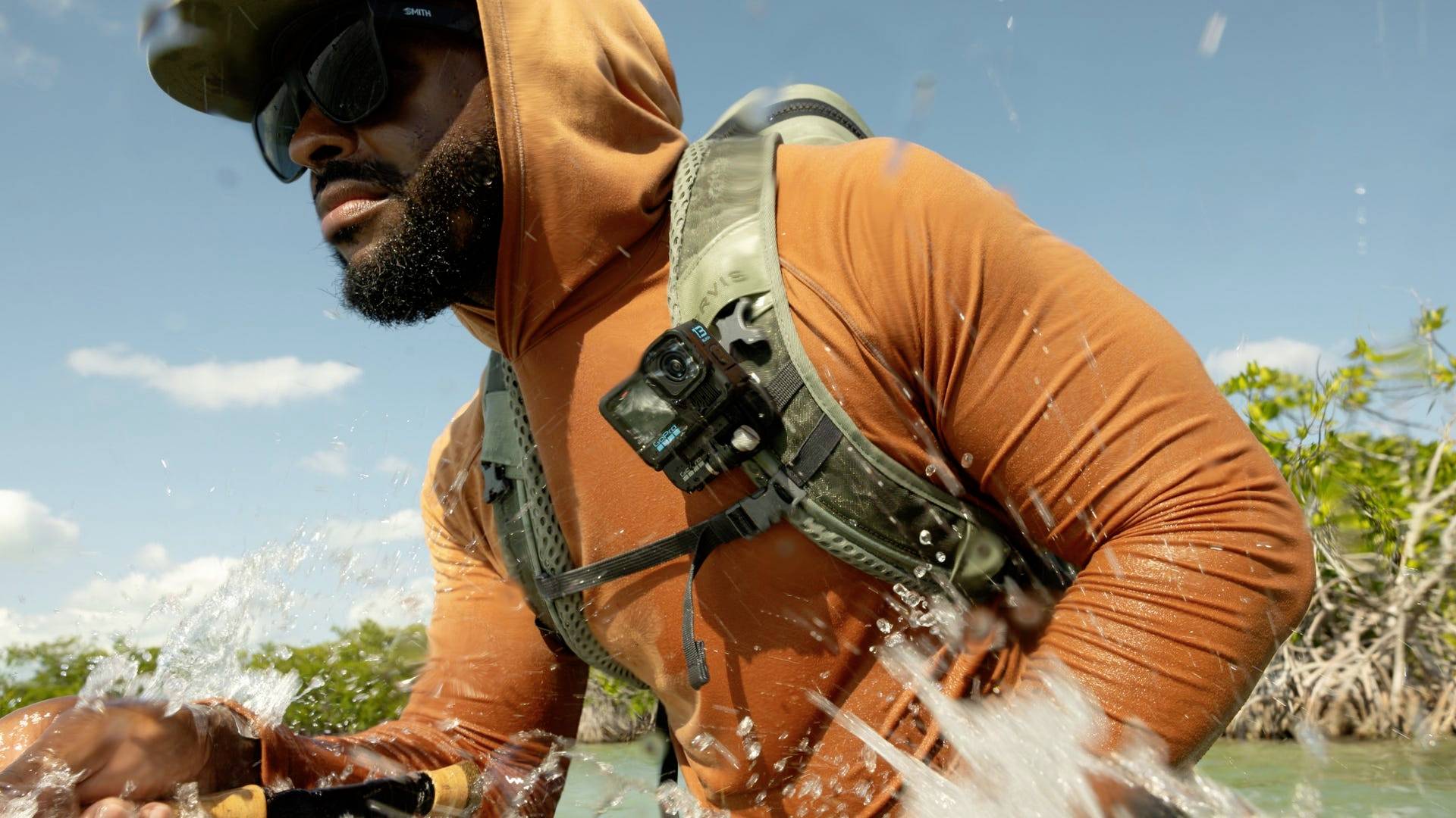 Une personne portant un sweat à capuche orange, des lunettes de soleil et un sac à dos, pataugeant dans de l'eau peu profonde. Le ciel est dégagé, avec des mangroves en arrière-plan.