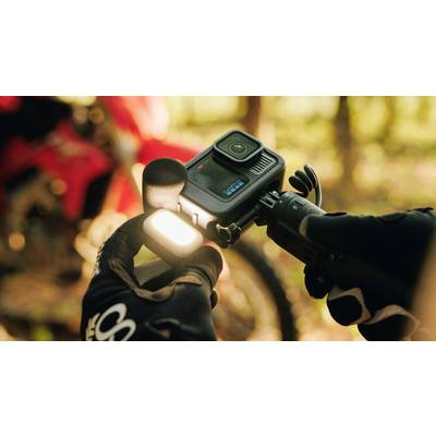 Une personne portant des gants noirs tient une caméra d'action et un microphone. À l'arrière-plan, une moto rouge floue est visible.