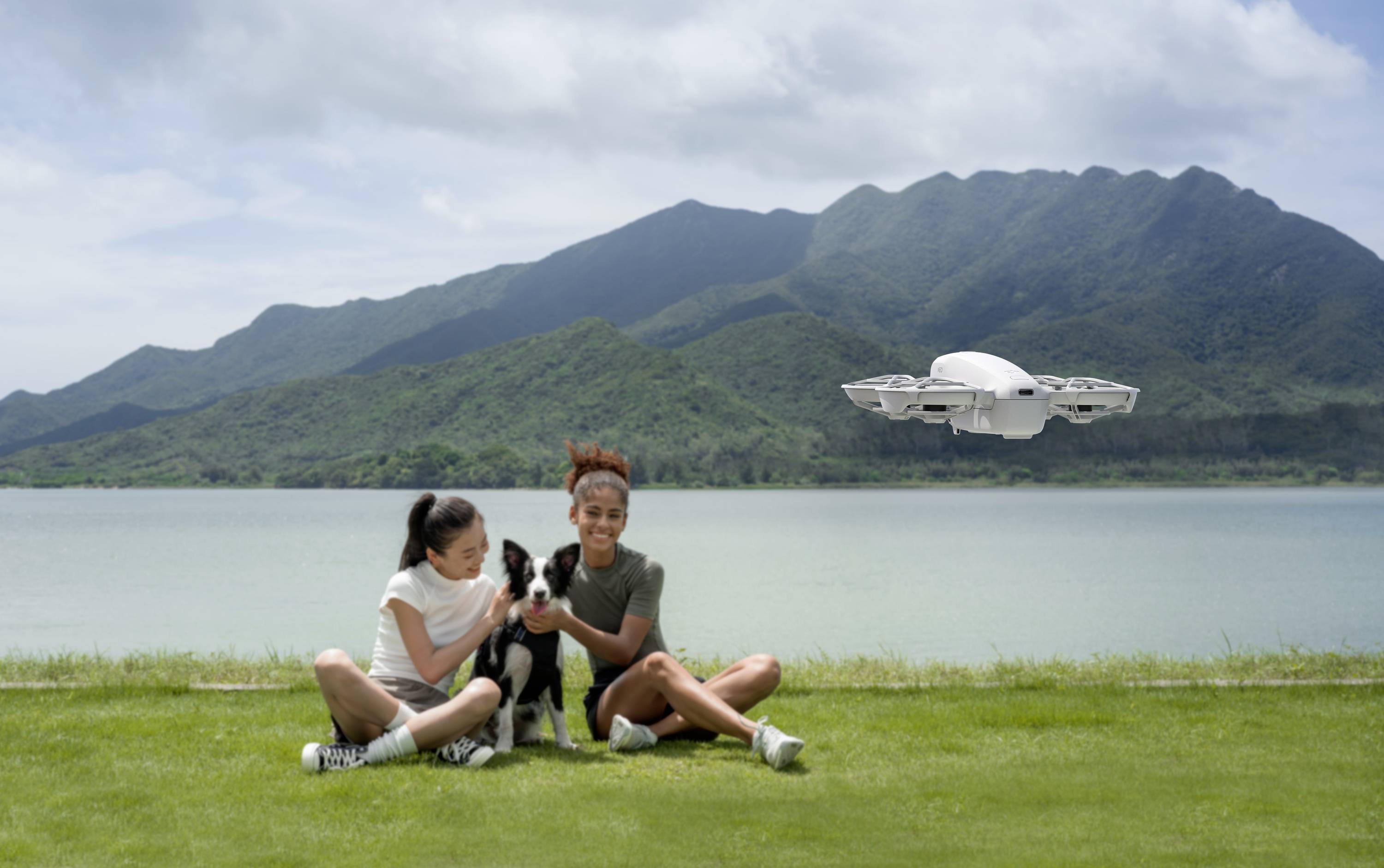 Deux personnes sont assises sur une prairie au bord d'un lac avec un chien. Un drone survole le premier plan. Des montagnes apparaissent en arrière-plan.