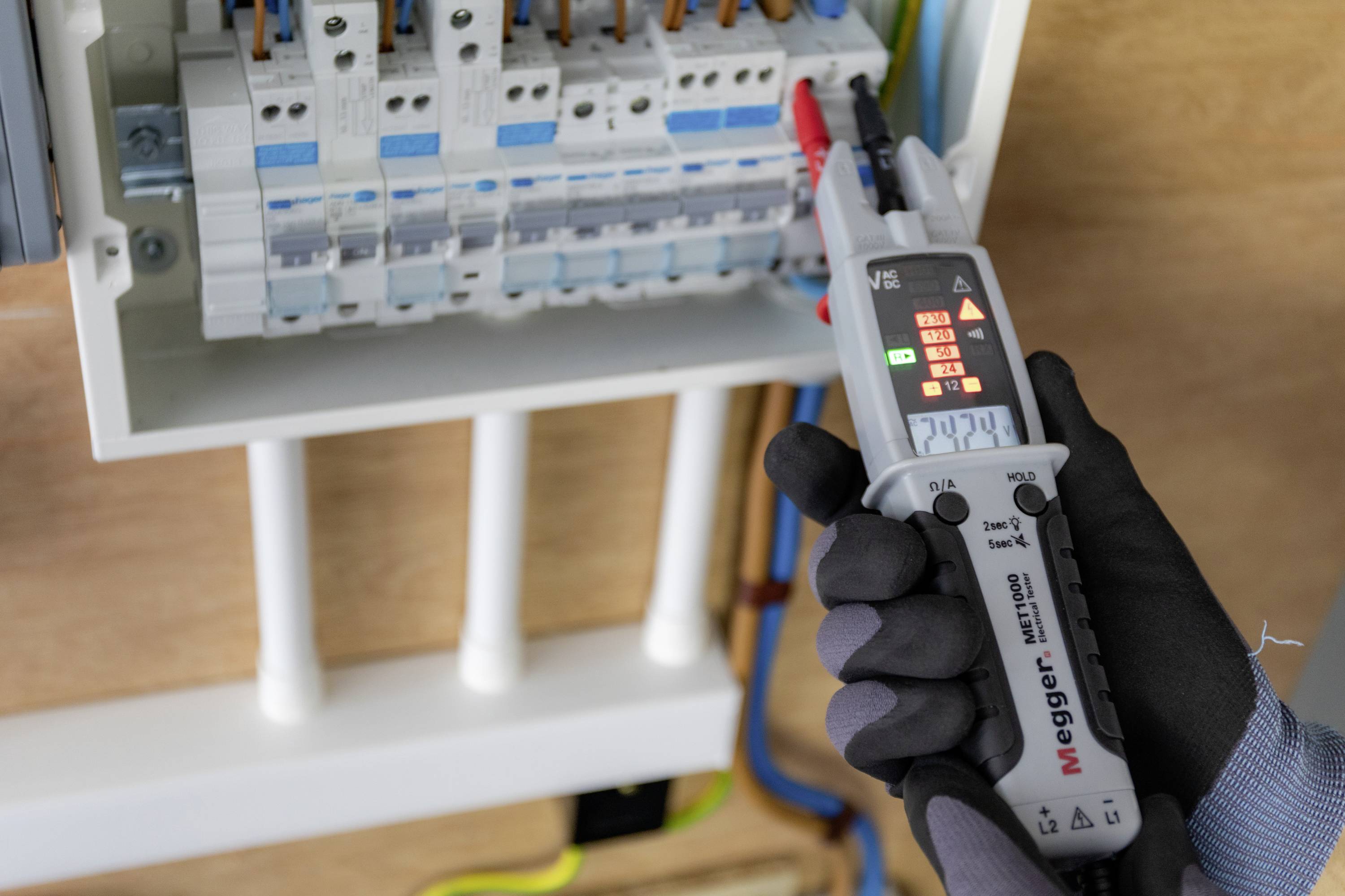 Un électricien vérifie un tableau électrique à l'aide d'un testeur de tension. Des gants le protègent des chocs électriques. L'affichage indique 234V.