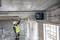 Un ouvrier du bâtiment portant un casque et un gilet haute visibilité mesure une distance sur un plafond en béton à l'aide d'un outil de précision sur un chantier de construction.