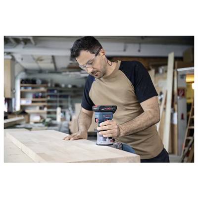 Un homme portant des lunettes de sécurité utilise un ponçoir électrique sur une surface en bois dans un atelier de menuiserie, concentré attentivement sur sa tâche.