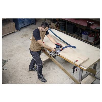 Une personne utilise un outil électrique pour travailler sur une grande pièce de bois dans un atelier. Des établis et des outils sont visibles en arrière-plan.