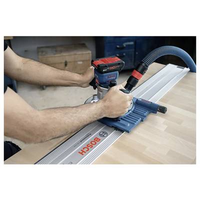 Une personne utilise un routeur Bosch sur une surface en bois, guidé par une voie métallique. L'espace de travail présente des outils et des tiroirs en arrière-plan.