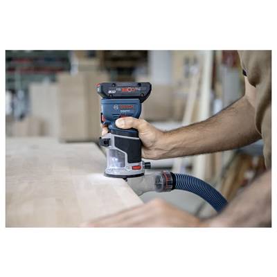 Une personne utilisant un outil de travail du bois Bosch sur une surface en bois, avec un tuyau de collecte de poussière attaché, dans un environnement d'atelier.