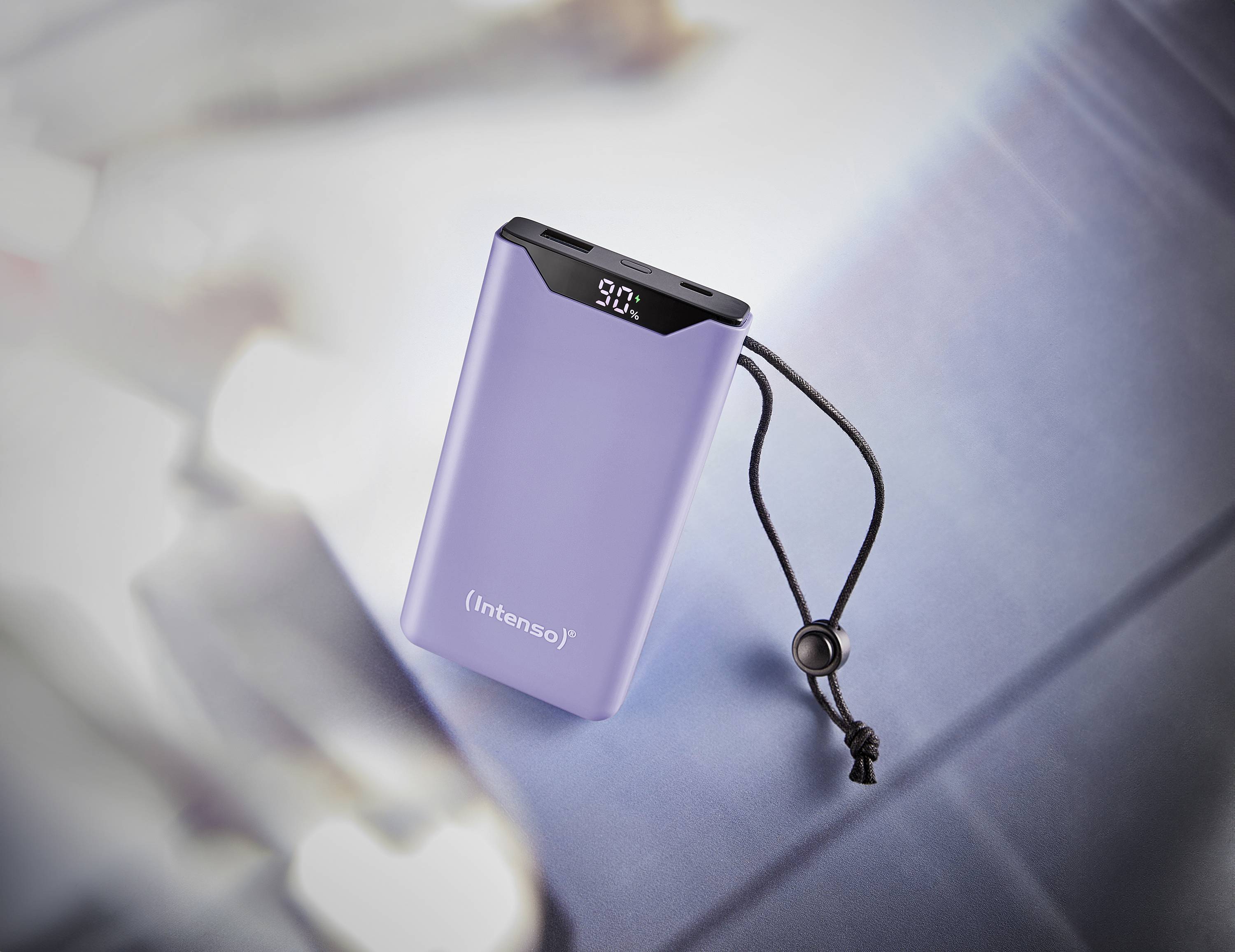 Powerbank Lila avec affichage numérique montrant 90% de charge. Elle est dotée d'un cordon de transport noir et repose sur une surface avec un arrière-plan estompé.