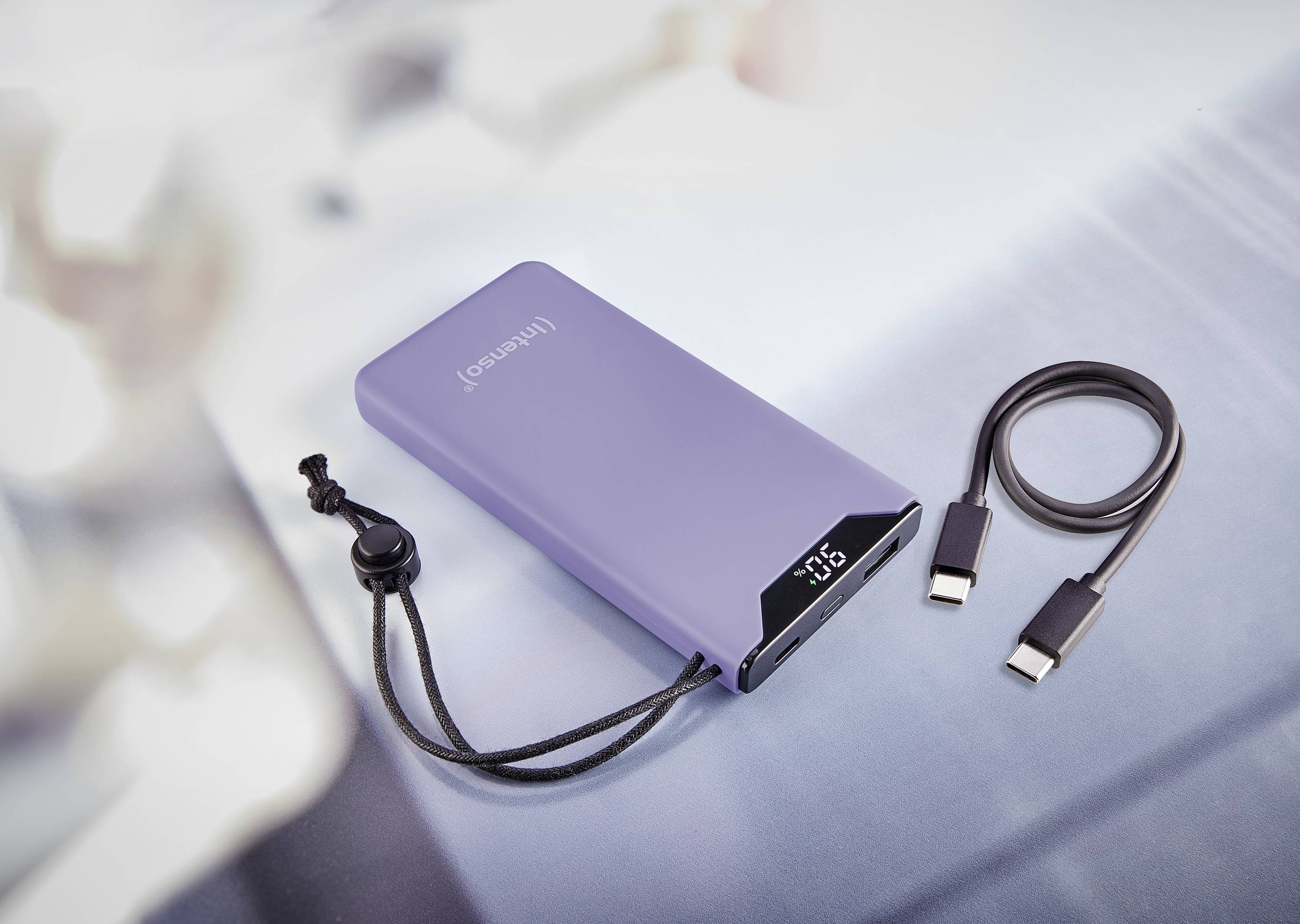 Batterie externe portable de couleur violette avec un petit écran LED affichant '100%'. Un câble USB est posé à côté.