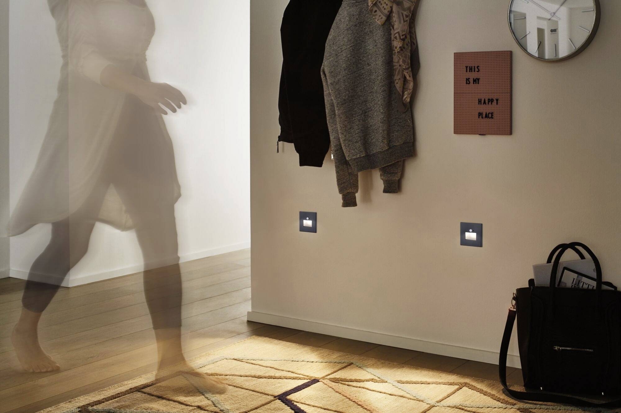 Une silhouette floue, immobile dans le couloir ; un miroir, des vestes et un panneau au mur. Le sol est éclairé par une lumière diffuse.